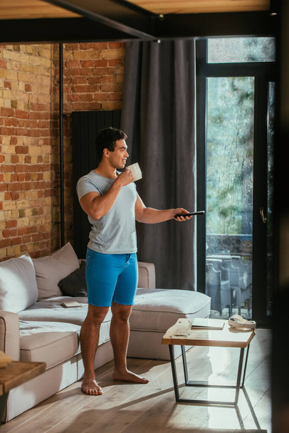 míchaný závod muž s šálkem kávy drží dálkové ovládání a sledování televize v obývacím pokoji během vlastní izolace - Fotografie, Obrázek