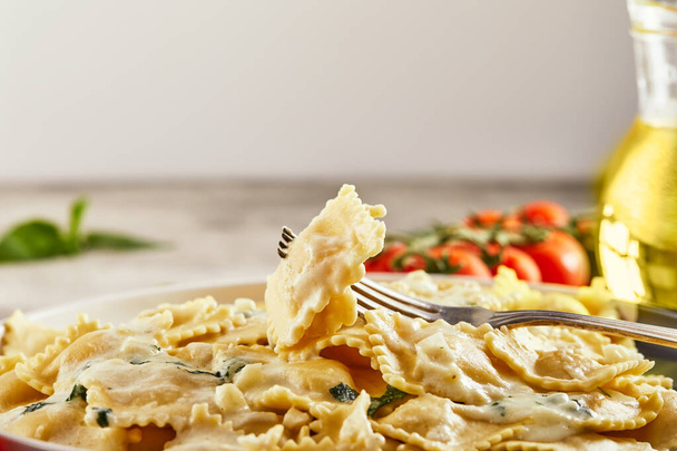 Tasty cooked ravioli with cream sauce, cherry tomatoes, sunflower oil and basil on a light wooden background. Copy space. Side view. - Photo, Image