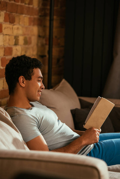 handsome smiling mixed race man reading book on sofa during quarantine - Foto, afbeelding
