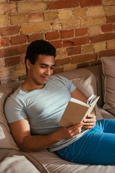 happy mixed race man reading book on sofa during quarantine - Fotoğraf, Görsel