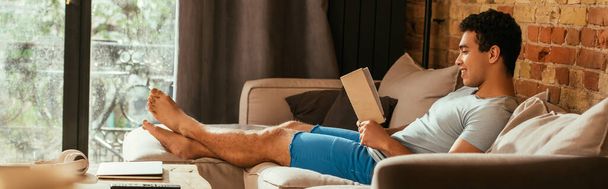 smiling mixed race man reading book on sofa during quarantine, website header - Foto, afbeelding