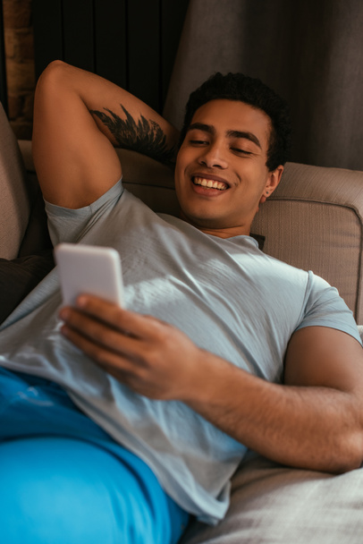 smiling mixed race man chilling and using smartphone on sofa during quarantine  - Fotografie, Obrázek