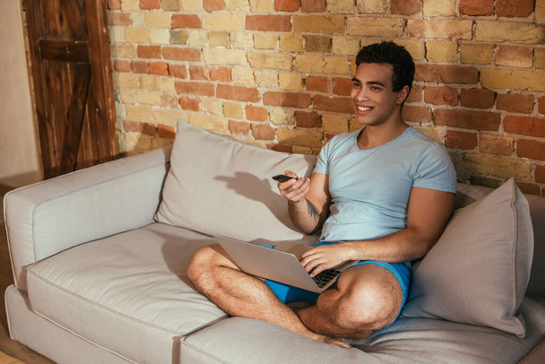homme de race mixte souriant avec ordinateur portable tenant télécommande et regarder la télévision dans le salon pendant la quarantaine
 - Photo, image