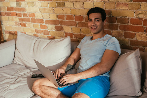 smiling bi-racial man chilling with laptop on sofa on quarantine - Фото, изображение