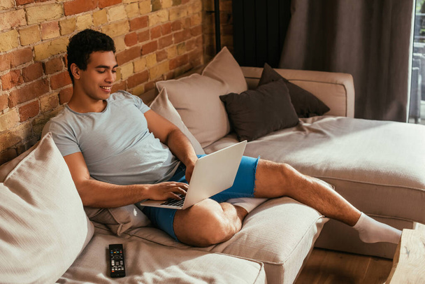 smiling mixed race man chilling with laptop on sofa with remote controller during self isolation - Foto, immagini