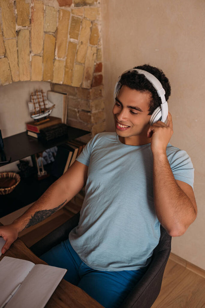 Lächelnder Mixed Race Freelancer, der während der Selbstisolierung im Home Office mit Kopfhörern und Notizblock arbeitet - Foto, Bild