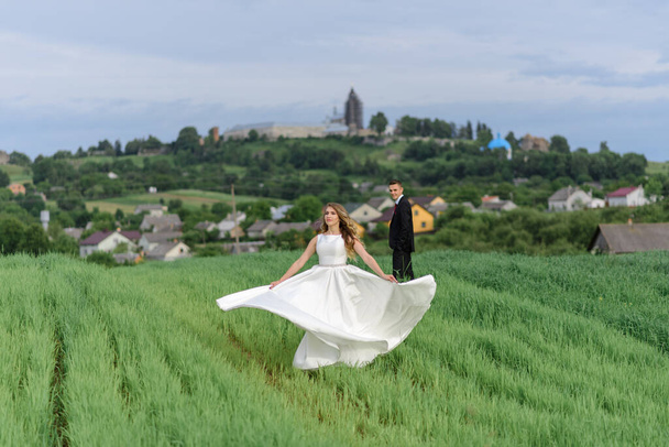 een echtpaar in een trouwjurk staat op een groen veld tegen de achtergrond van een dorp bij zonsondergang, de bruid en bruidegom - Foto, afbeelding