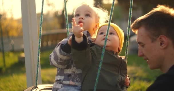 Slow motion: Happy young father pushing his sons on the swing. Father and two son playing at home on the yard with tire swing hanging from tree with beautiful sunlight. two brothers and father swing. - Footage, Video