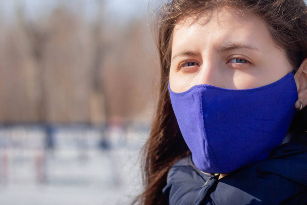 Portrait of a girl in a winter jacket with a hood and in a blue protective mask on her face strolling through a deserted amusement park. - Фото, изображение
