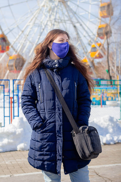 Ritratto di una ragazza in giacca invernale con cappuccio e maschera protettiva blu sul viso passeggiando in un parco divertimenti deserto
. - Foto, immagini