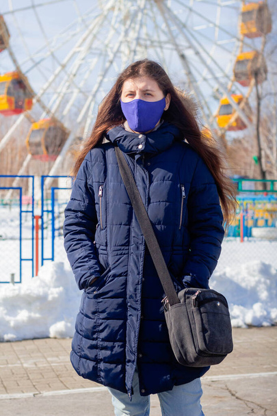 Portret dziewczyny w zimowej kurtce z kapturem i niebieską maską ochronną na twarzy spacerującej po opuszczonym parku rozrywki. - Zdjęcie, obraz