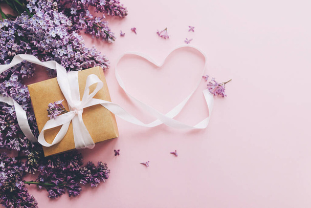 Lilac flowers, gift box and heart ribbon on pink paper, stylish flat lay with copy space. Floral greeting card. Happy mothers day concept. Purple lilac flowers bouquet with craft present box - Foto, Imagem