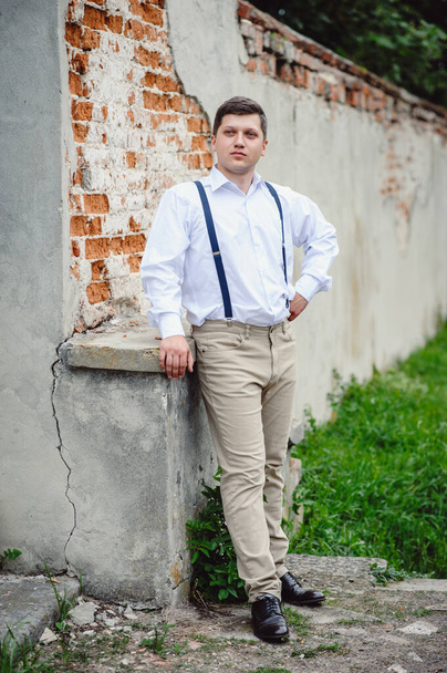 Привлекательный успешный молодой бизнесмен, одетый в белую рубашку и светлые брюки с подтяжками, стоит возле серой винтажной стены. Стиль и мода - Фото, изображение