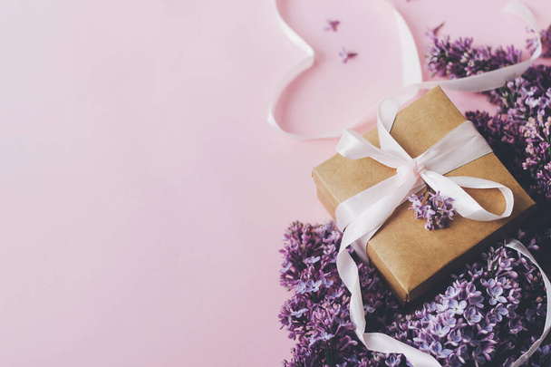 Сирень цветы, подарочная коробка и сердечная лента на розовой бумаге. Стильная цветочная открытка с копировальным пространством. Счастливого Дня Матери. Фиолетовый букет сиреневых цветов с коробкой подарков
 - Фото, изображение