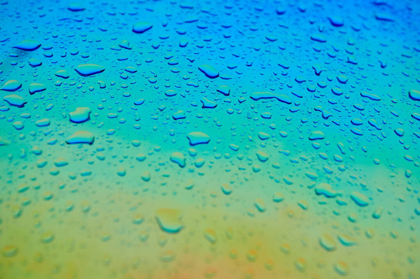 Σταγόνες νερού στην ολογραφική επιφάνεια χαμαιλέοντα. Όμορφο πολύχρωμο φόντο με ρηχό βάθος πεδίου. Μπλε και κίτρινη κλίση. - Φωτογραφία, εικόνα