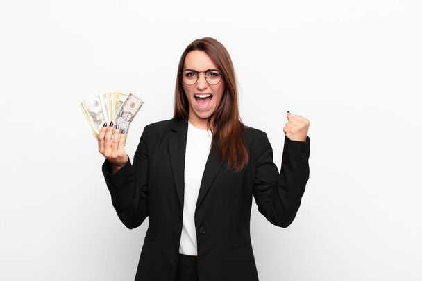 young businesswoman shouting aggressively with an angry expression or with fists clenched celebrating success with dollar banknotes - Фото, изображение