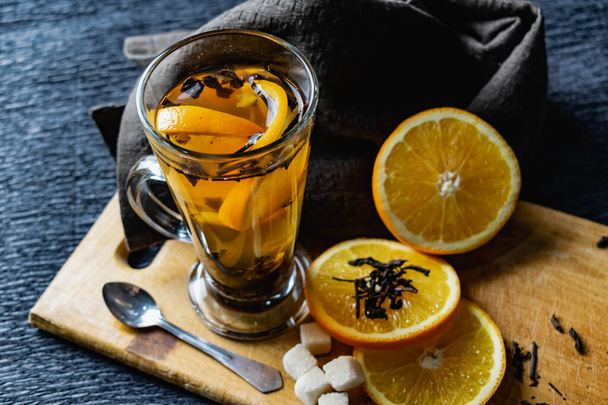 Citrus drink, tea with petals in a transparent glass mug with slices of sweet orange and refined sugar. Slicing fruit on a wooden Board with a brown napkin in a cozy dark background in a low key - Фото, изображение