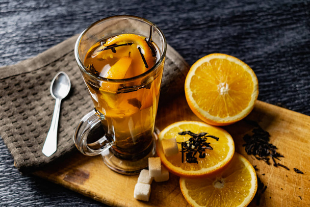 Citrus drink, tea with petals in a transparent glass mug with slices of sweet orange and refined sugar. Slicing fruit on a wooden Board with a brown napkin in a cozy dark background in a low key - 写真・画像