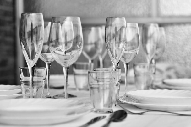 Close up empty clean diverse of glassware and porcelain plates served on white tablecloth, no people. Table settings wait for guests at home or restaurant. Dinner party, tableware shop ad, bw image concept - Фото, изображение