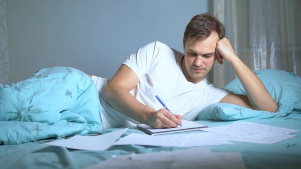 A man writes on paper documents lying in bed. work at home. - Фото, изображение