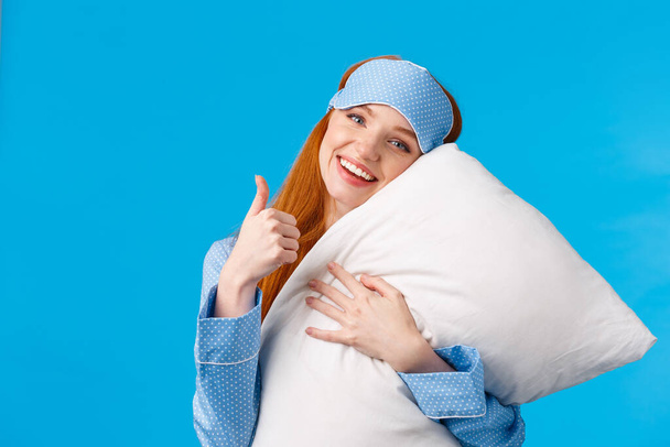 Saying yes to good healthy sleep. Cheerful pretty redhead woman in sleep mask and nightwear, hugging soft pillow showing thumbs-up in approval, like gesture, standing pleased blue background - Zdjęcie, obraz