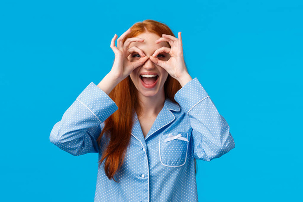 Amused girl seeing wonderful promo, want try out. Attractive redhead woman in lovely pyjama make glasses with fingers, looking through smiling thrilled and excited, standing in nightwear - Fotó, kép