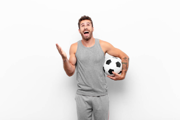 young handsome man feeling happy, amazed, lucky and surprised, celebrating victory with both hands up in the air holding a soccer ball - Photo, image