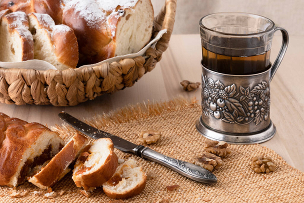 Нарезанный свежий хлеб и винтажный нож. Чай в стакане с держателем для чашки. Крошки хлеба. Светлый деревянный фон. Вид сбоку. Крупный план
 - Фото, изображение