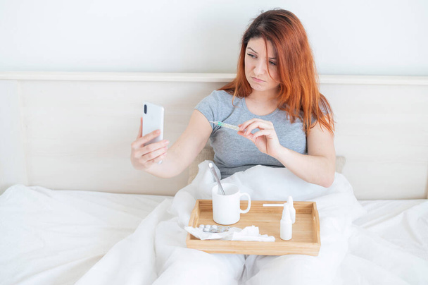 A cold blogger is recording a video for his vlog. A woman with a fever sits in bed and shows a thermometer to the smartphone camera. The girl is sick and takes a selfie. - Foto, Imagen
