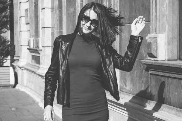 Mulher morena com um sorriso positivo em um vestido e couro casaco preto agitar o cabelo na rua e desfruta de um dia de primavera ensolarado. Viagem de menina em Batumi, Geórgia. Cidade velha no fundo. - Foto, Imagem