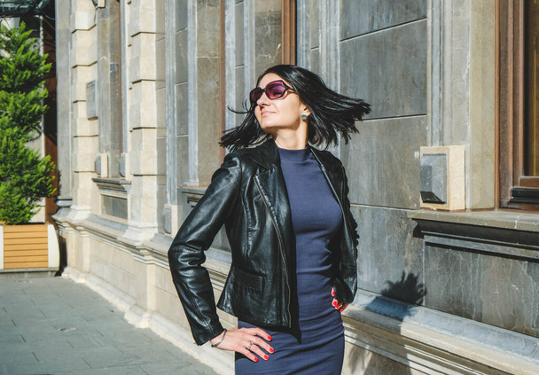 Brunette woman with a positive smile in a dress and leather black jacket shake hair in street and enjoys a sunny spring day. Girl travel in Batumi, Georgia. Old city at background. - Fotografie, Obrázek