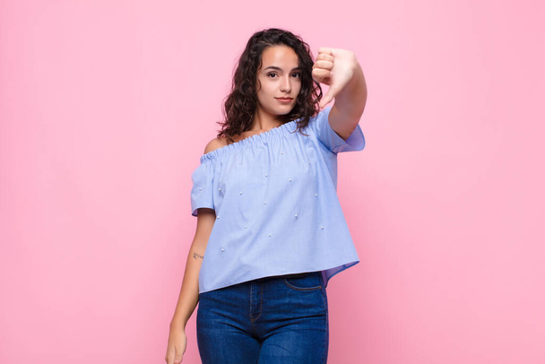 young pretty woman feeling cross, angry, annoyed, disappointed or displeased, showing thumbs down with a serious look against pink wall - Fotoğraf, Görsel