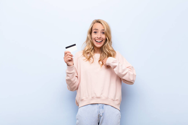 young blonde woman feeling happy, surprised and proud, pointing to self with an excited, amazed look holding a credit card - Fotografie, Obrázek
