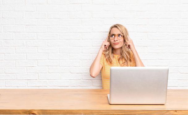 young blonde woman feeling confused or doubting, concentrating on an idea, thinking hard, looking to copy space on side using a laptop - Foto, Bild