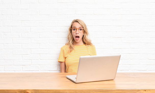 young blonde woman looking very shocked or surprised, staring with open mouth saying wow using a laptop - Foto, Imagem