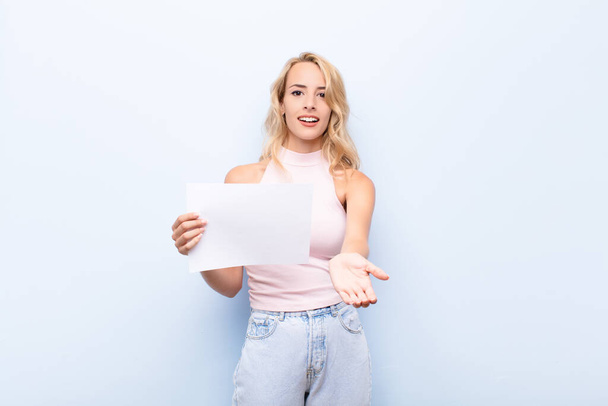 young blonde woman smiling happily with friendly, confident, positive look, offering and showing an object or concept holding a sheet of paper - Photo, image