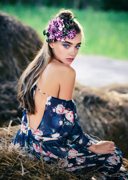 Romantic cute girl sitting on the hay in a dress with an open back. A young girl near a haystack with a flower on her head. Looks into the frame, outdoor portrait - Foto, immagini