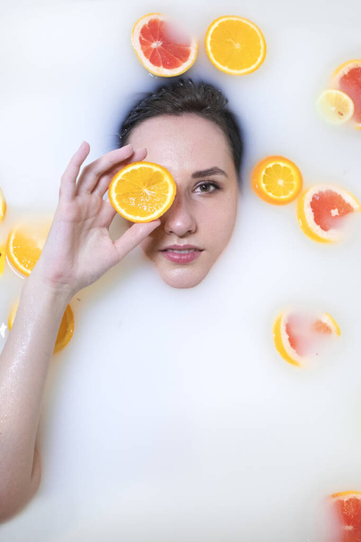 Woman portrait in milk bath with oranges, lemons and grapefruits. Healthy dewy skin. Fashion model girl, spa and skin care concept. Spring colours - yellow, orange, red. - Photo, Image