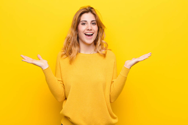 young pretty blonde woman feeling happy, excited, surprised or shocked, smiling and astonished at something unbelievable against yellow wall - Zdjęcie, obraz