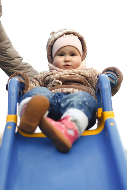 A happy child sits on a children's slide and smiles in a stylish brown jacket and hat in autumn. Fashion and style for children. - Foto, Imagen