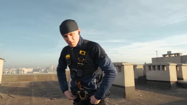 Sportowanie europejskich mężczyzn wspinacza przemysłowego w mundurze przymocowuje różne haki i linki do jego ciała, stojąc na dachu drapacza chmur. Ekstremalnie niebezpieczna praca związana z wysokością. Ryzyko dla życia - Materiał filmowy, wideo