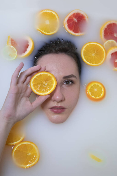 Vrouwenportret in melkbad met sinaasappels, citroenen en grapefruits. Gezonde dauwhuid. Mode model meisje, spa en huidverzorging concept. Voorjaarskleuren - geel, oranje, rood. - Foto, afbeelding