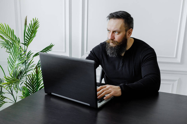a bearded man in black golf works at a laptop at home in a bright room. Self isolation - Zdjęcie, obraz