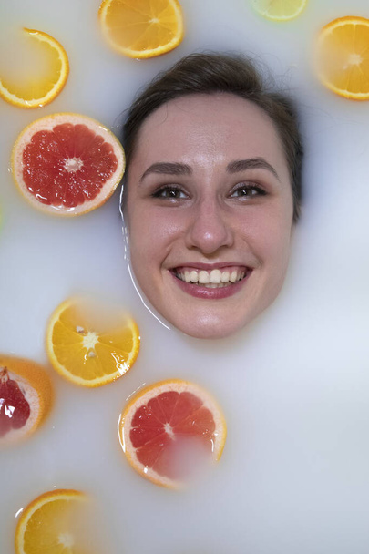 Woman portrait in milk bath with oranges, lemons and grapefruits. Healthy dewy skin. Fashion model girl, spa and skin care concept. Spring colours - yellow, orange, red. - Photo, image