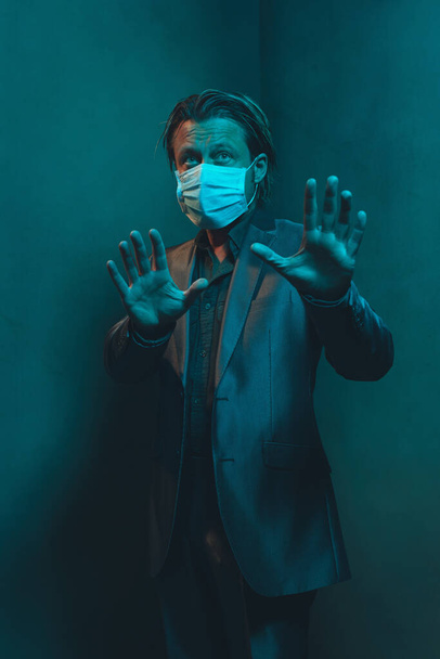 Homme avec masque hygiénique en costume se tient dans le coin de la pièce vide. Faire un geste de distance de maintien avec les mains
. - Photo, image