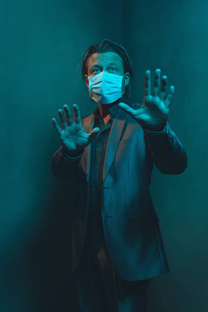 Man with hygienic face mask in suit stands in corner of empty room. Making holding distance gesture with hands. - Photo, Image