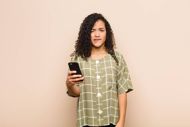 young black woman looking puzzled and confused, biting lip with a nervous gesture, not knowing the answer to the problem with a smartphone - Foto, Imagem
