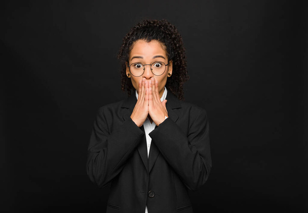 young black woman happy and excited, surprised and amazed covering mouth with hands, giggling with a cute expression against black wall - 写真・画像