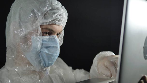 wetenschapper arts man in wit pak en masker voorzichtig opent een laptop op een zwarte achtergrond close-up - Foto, afbeelding