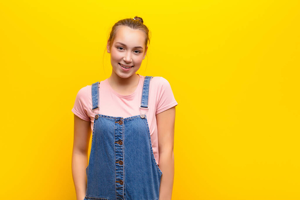 young blonde pretty girl with a big, friendly, carefree smile, looking positive, relaxed and happy, chilling against yellow wall - Foto, imagen